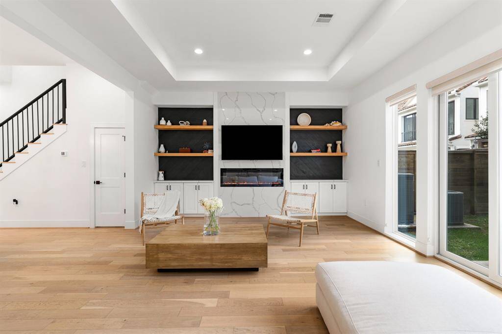 This modern living room features sleek hardwood floors, large windows for natural light, a wall-mounted TV above a contemporary fireplace, built-in shelving, and an open staircase with black railing.