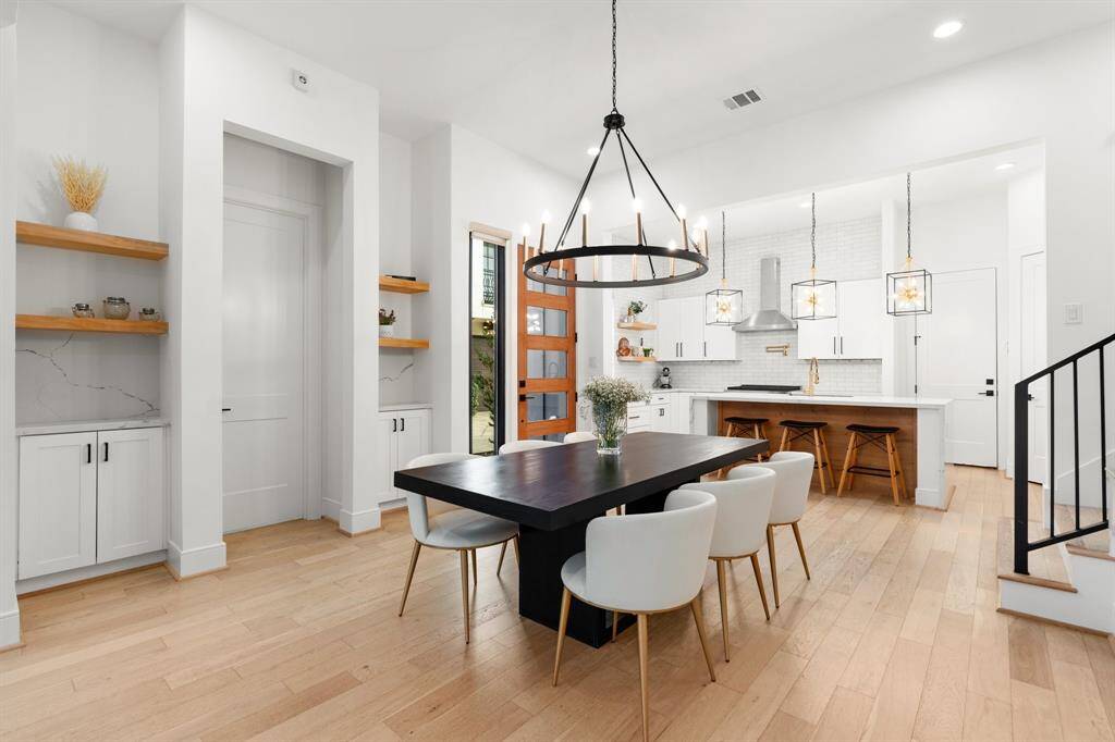 This home showcases a modern, open-concept dining and kitchen area. It features light wood flooring, white cabinetry. The kitchen includes a central island, contemporary pendant lighting, and stainless steel appliances. The space is bright with a minimalist design, offering a welcoming and stylish atmosphere.