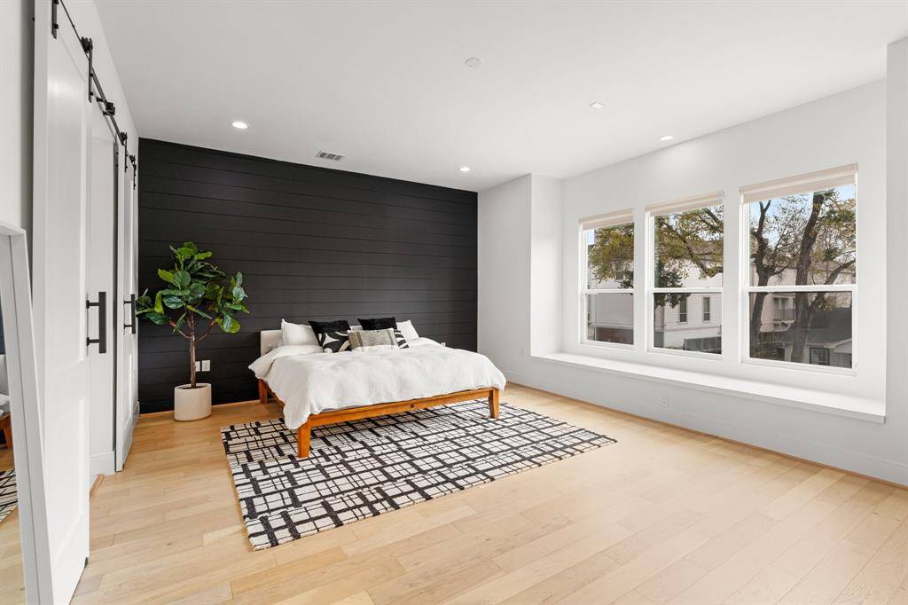 This spacious bedroom features a modern design with a striking black accent wall, large windows offering plenty of natural light, and light wood flooring. Complemented by a sliding barn door leading to the en-suite bathroom