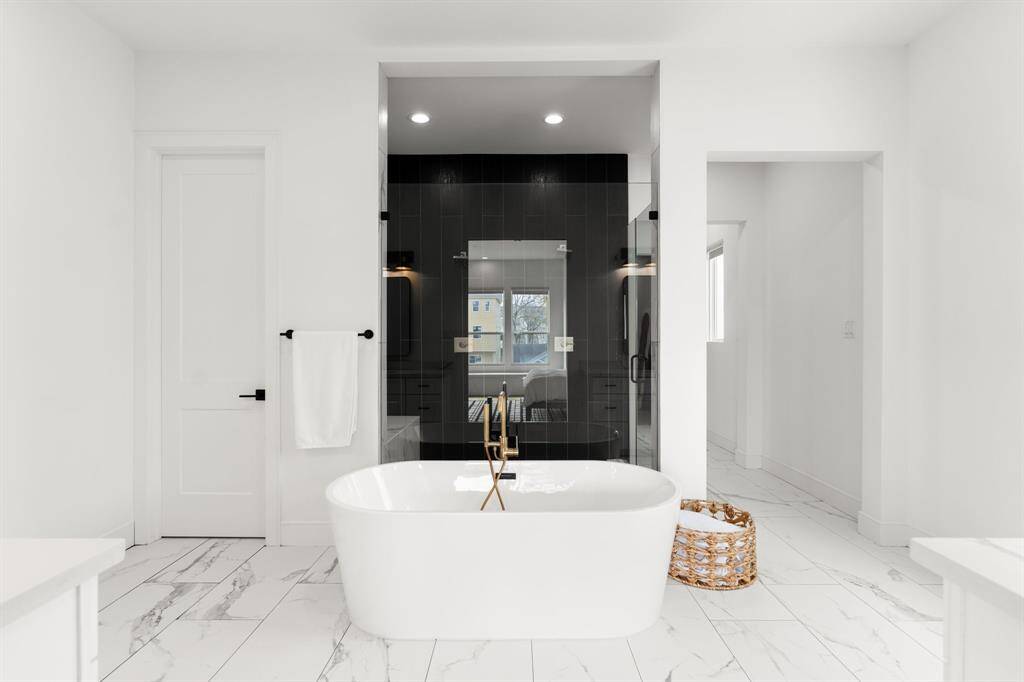 This bathroom features a modern, freestanding tub with a sleek black accent wall and glass-enclosed shower. The space is bright and spacious with white marble-style flooring, creating a luxurious, spa-like atmosphere.