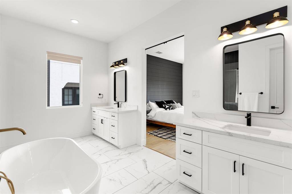 This bathroom features a modern design with white cabinetry, countertops, and a freestanding tub. It has dual vanities with stylish lighting and a window for natural light. The space connects to a bedroom with a sleek, dark accent wall.
