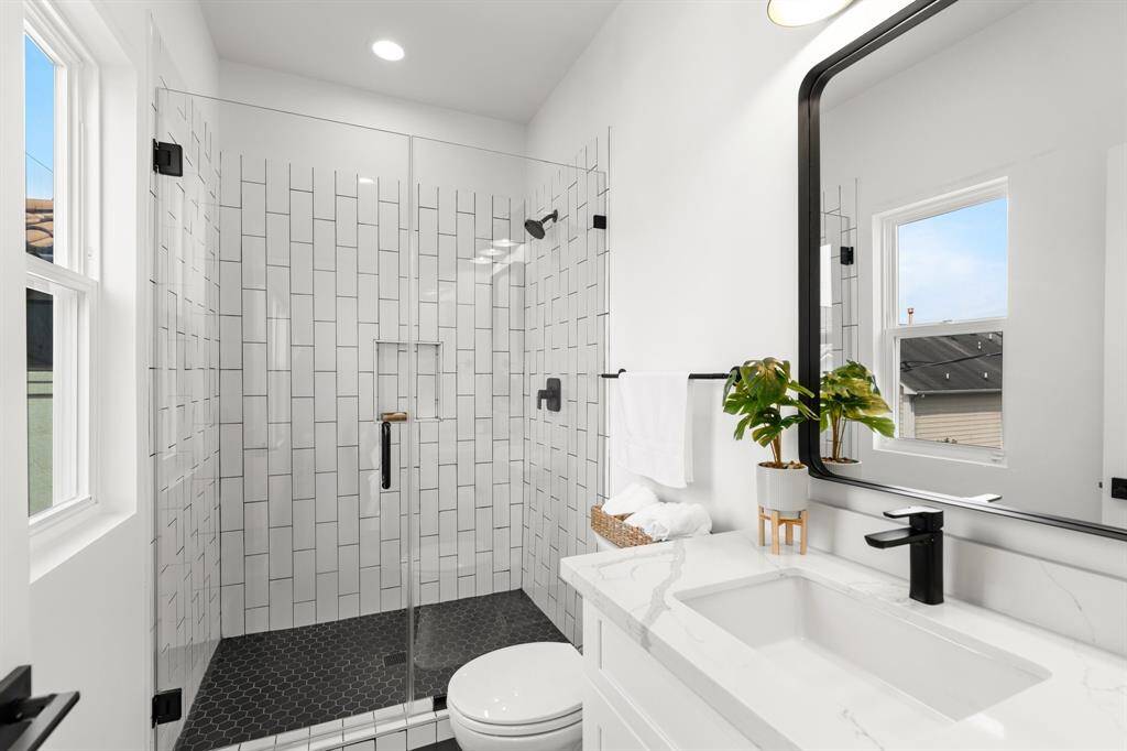 This bathroom features a modern design with a glass-enclosed shower, white subway tiles, and a sleek black showerhead. The vanity includes black fixtures, and a large mirror. Natural light flows in through a window, complementing the bright, clean aesthetic.