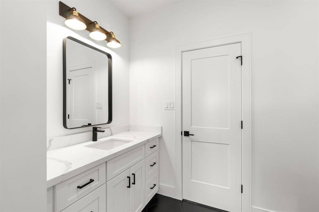 This bathroom features a modern design with a white vanity, black fixtures, and a sleek mirror. The space is well-lit with three overhead lights, and the overall aesthetic is clean and minimalist.