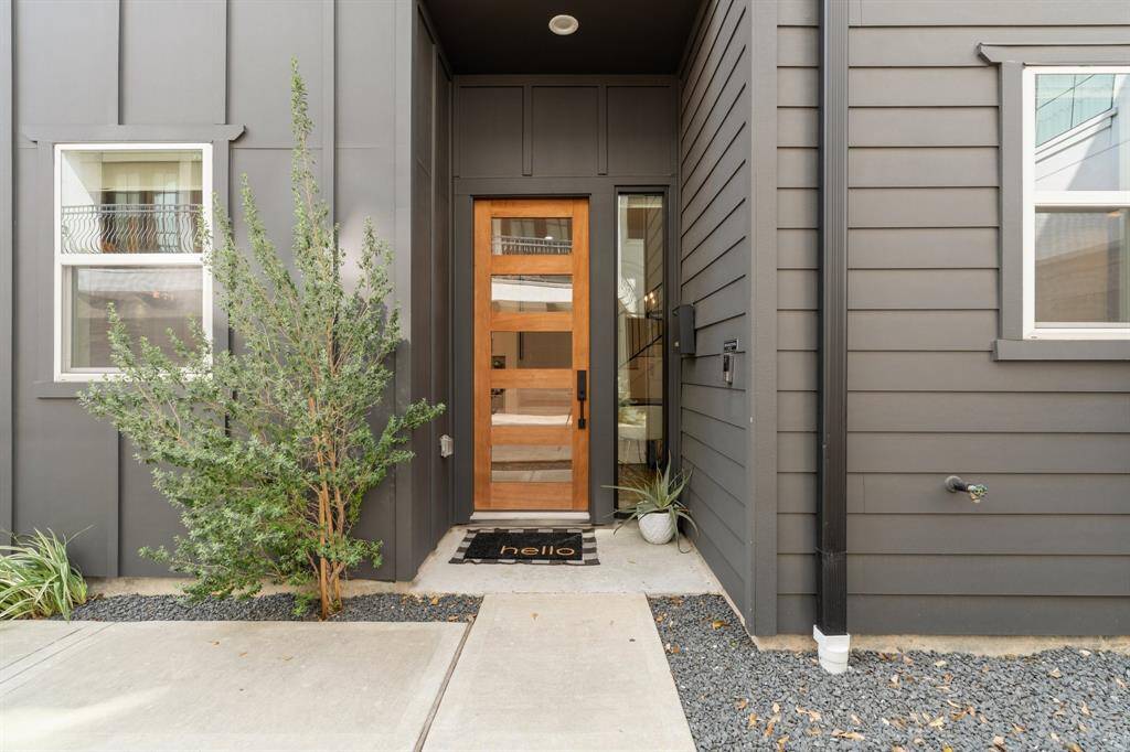 This modern entryway features a sleek wooden door with glass panels, surrounded by dark gray siding. The space is accented with minimalist landscaping, including a small tree and potted plants, creating a welcoming and stylish front entrance.
