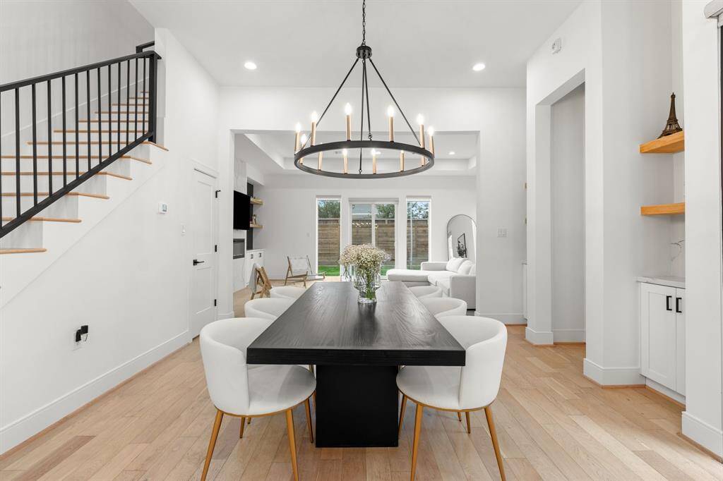 This modern living space features an open floor plan with light wood flooring and a striking black staircase. A sleek dining are is complemented with a contemporary chandelier. The room flows into a cozy seating area with large windows, offering ample natural light.