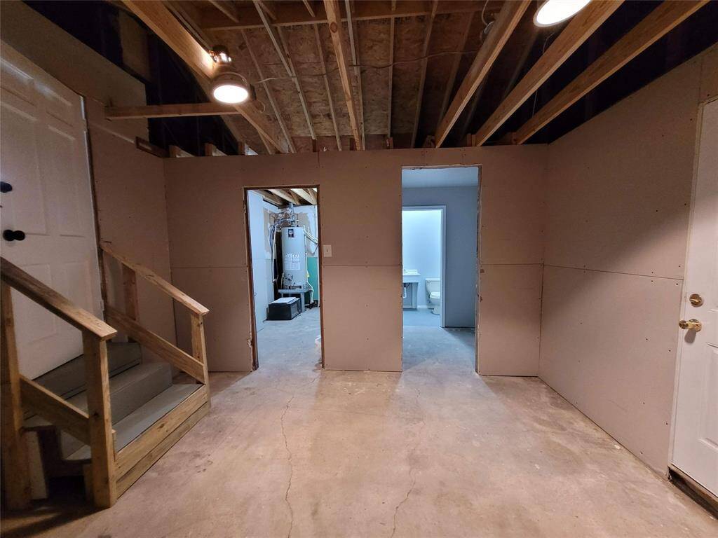 A partially finished garage with exposed wooden ceiling beams, drywall, and concrete flooring. It features a staircase, doors leading to utility and bathroom areas, and overhead lighting, offering potential for customization.