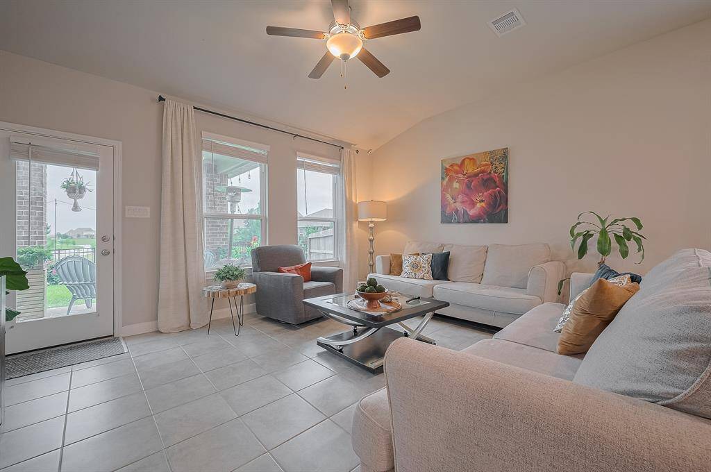 Bright and welcoming living room with neutral tones, tiled flooring, and a ceiling fan, features large windows for natural light. Access to a patio adds indoor-outdoor appeal.