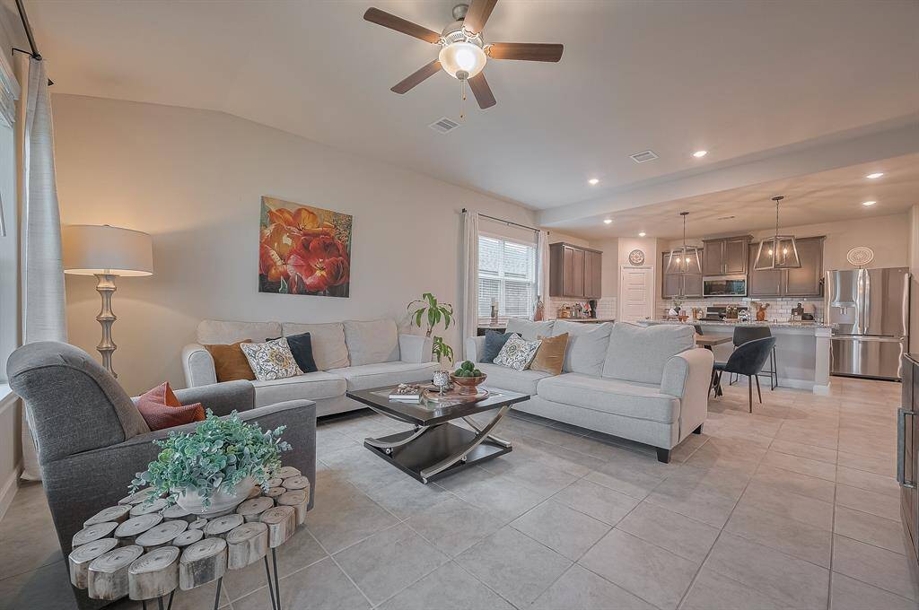 The room flows seamlessly into a contemporary kitchen equipped with stainless steel appliances and stylish pendant lighting. A ceiling fan and large windows ensure a bright and airy atmosphere.