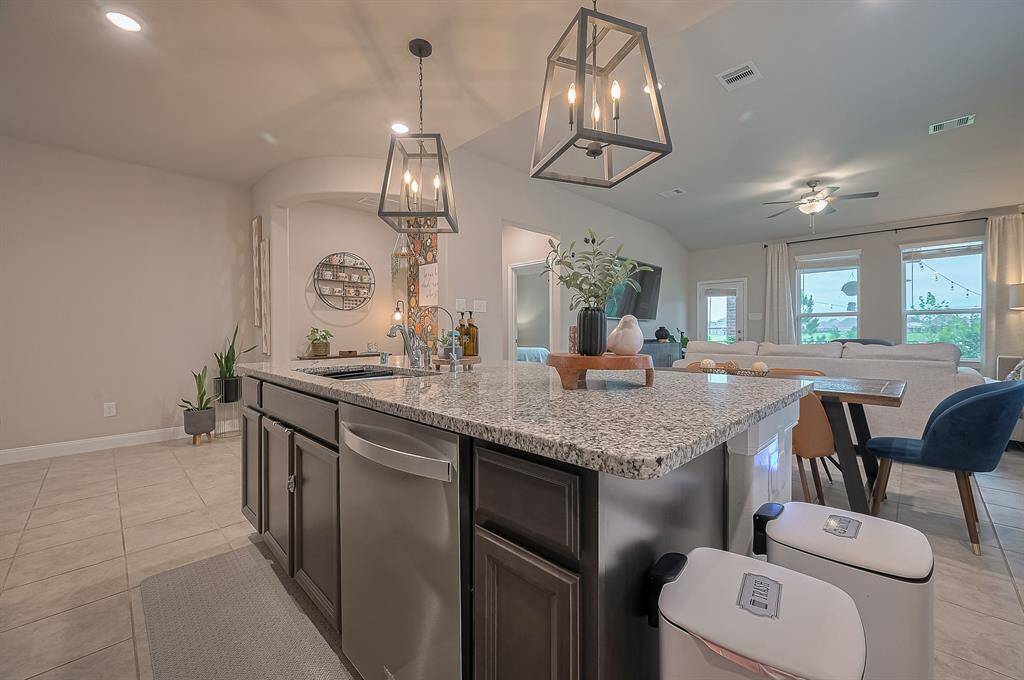 The kitchen features a large granite island with under-counter storage, pendant lighting, and stainless steel appliances. The space flows seamlessly into a cozy living room with ample natural light from multiple windows, neutral tones, and stylish decor.