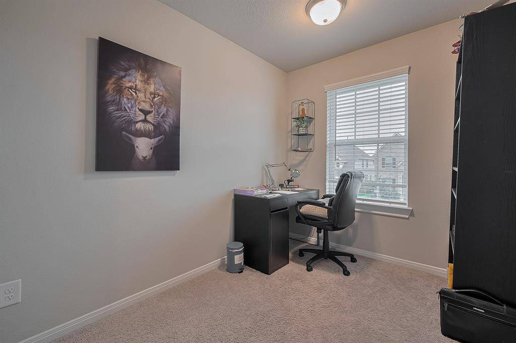 This photo shows a cozy home office with carpeted flooring and neutral walls. It features a window with blinds providing natural light.