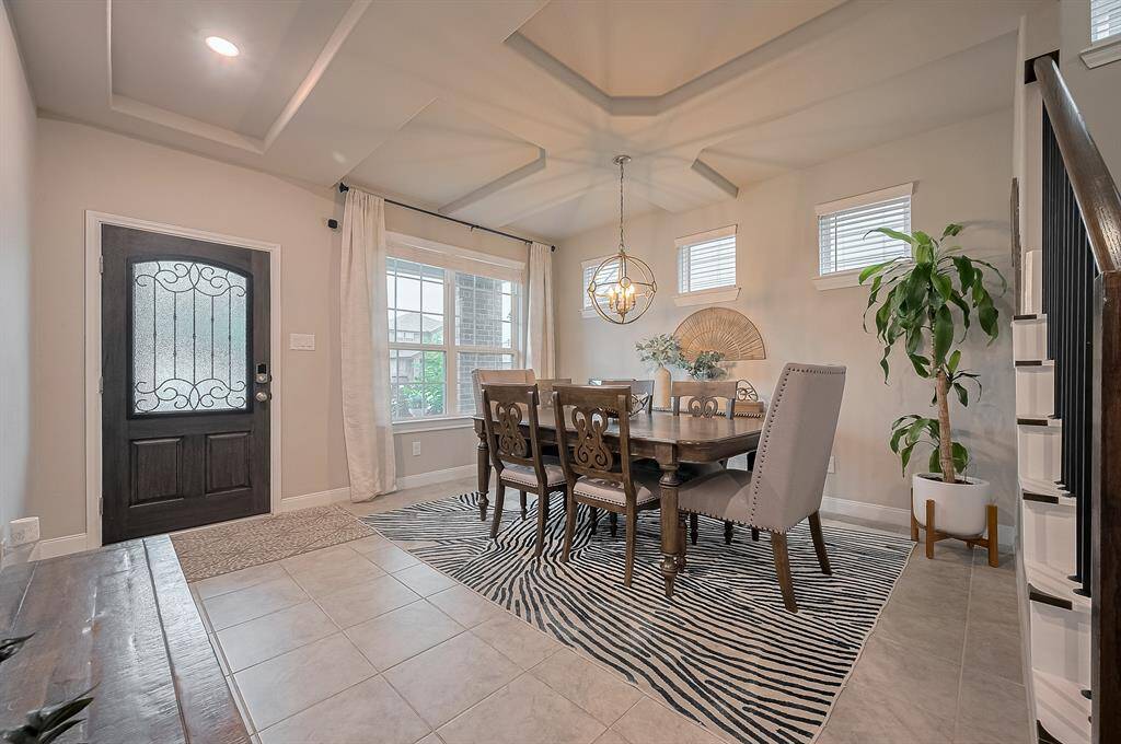 This welcoming entryway and dining area features elegant tile flooring, a stylish dark wood door with ornate glass, and ample natural light from large windows. The coffered ceiling and modern chandelier add sophistication.