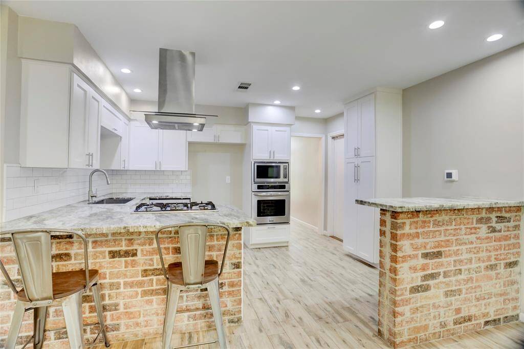 A look into the rustic kitchen from the living room. Kitchen includes a gas cooktop, stainless steel sink, oven, and microwave, along with a breakfast bar and separate standing bartop that could be used as a buffet, dry bar, or additional seating.