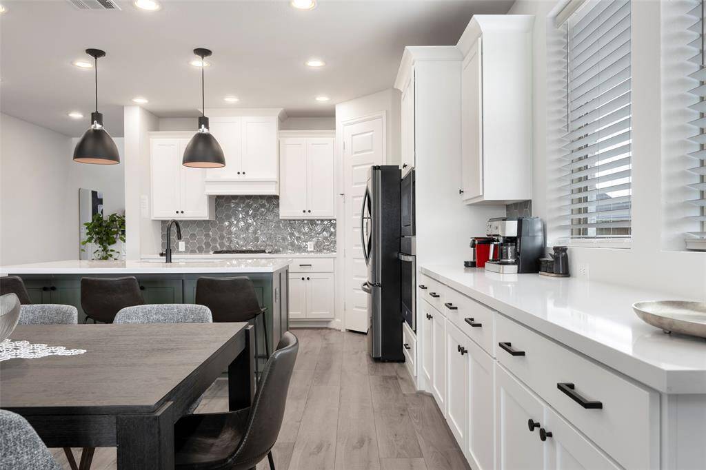 This kitchen features a modern design with sleek white cabinetry, a stylish hexagonal backsplash, and black appliances. It includes a spacious island with pendant lighting, an adjacent dining area, and ample natural light through large windows, creating a bright and inviting space.