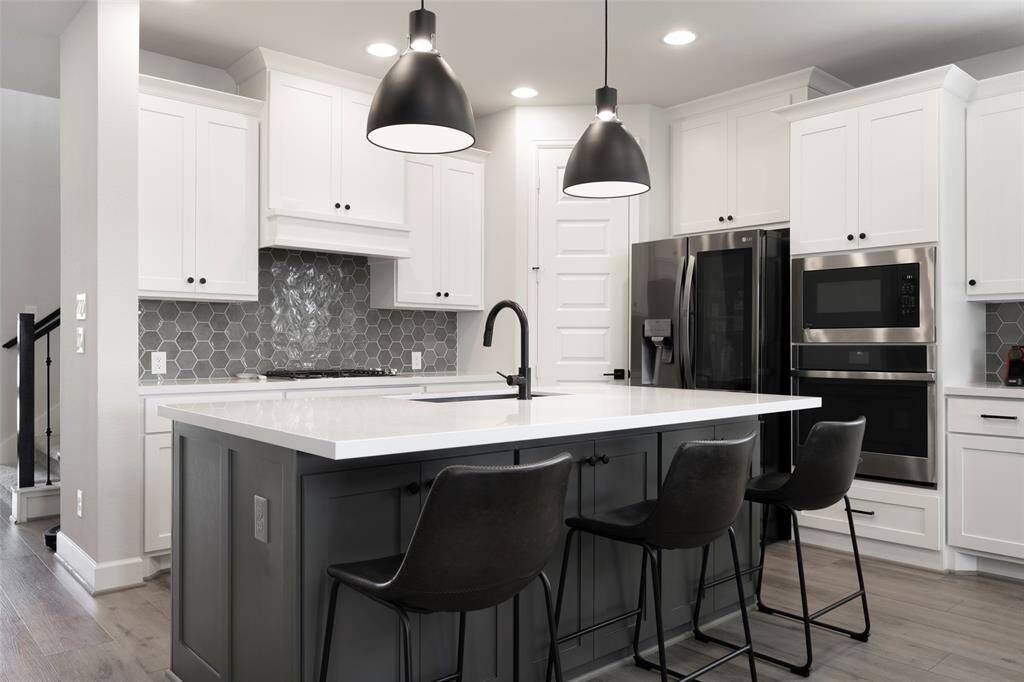 This modern kitchen features a sleek design with white cabinetry, a large island with a waterfall countertop, and contemporary black pendant lighting. It includes stainless steel appliances, a hexagonal tile backsplash, and an open concept layout with bar seating.