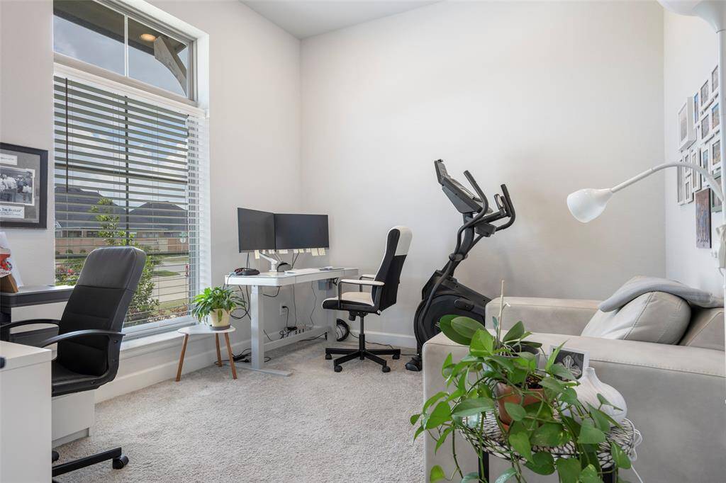 Bright and versatile home office featuring a large window, modern desk setup, room for exercise equipment, and cozy seating.