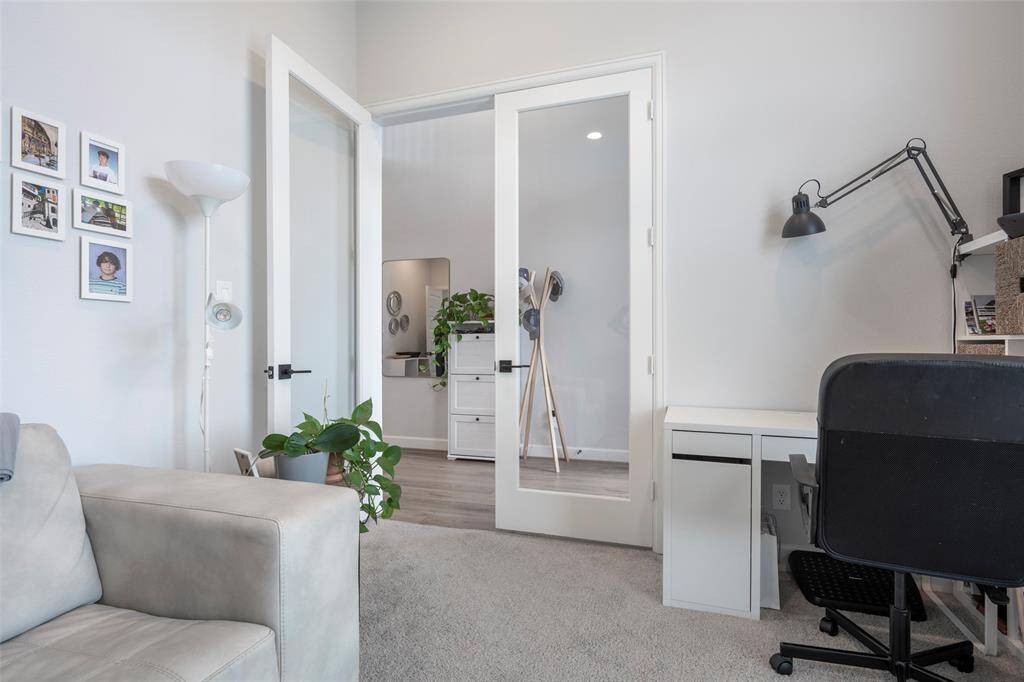 This photo showcases a bright, modern office space with a glass-paneled door, neutral walls, and carpeted flooring. It features a comfortable seating area, a sleek desk with a lamp, and lively green plants, creating an inviting and functional work environment.