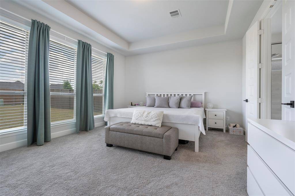 This spacious bedroom features large windows with elegant drapes, allowing plenty of natural light. It includes a comfortable bed with a cushioned bench at the foot and is complemented by neutral tones and plush carpeting.