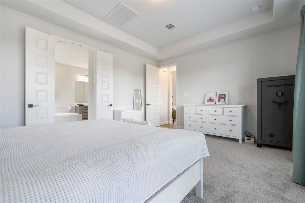 This bright bedroom features a neutral palette, plush carpeting, and tray ceiling, an en-suite bathroom through double doors.