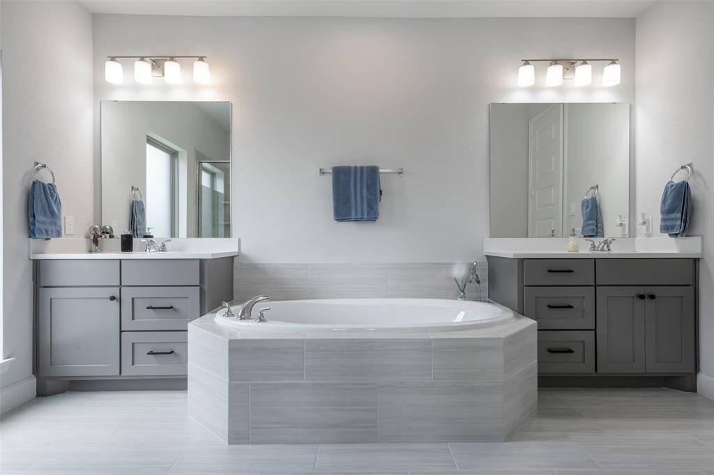 This photo showcases a modern bathroom with a spacious, central bathtub, dual vanities, ample lighting, and sleek gray cabinetry, creating a clean and inviting atmosphere.