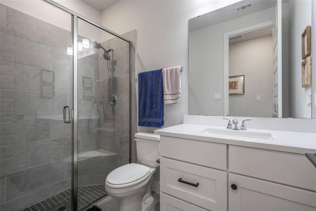 Modern bathroom with a glass-enclosed shower featuring gray tiles. It includes a sleek white vanity with a large mirror, under-sink storage, and a toilet. Bright and clean design.