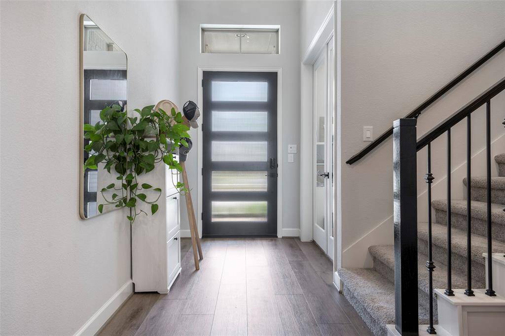 This photo showcases a modern, bright entryway with a sleek frosted glass front door, wood flooring, and a carpeted staircase with black railing. A large wall mirror and a green plant add a touch of elegance and freshness.