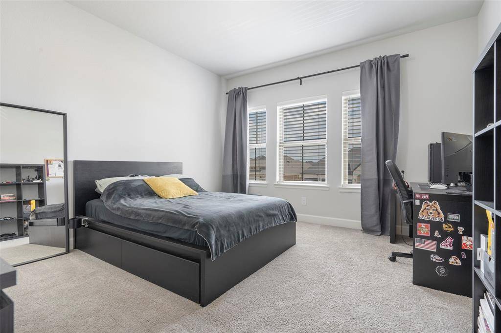 A bright, spacious bedroom featuring a large bed with a dark frame, neutral carpet, and ample natural light from three windows with gray curtains. The room includes a desk with a computer, and a mirror, creating a modern and functional space.