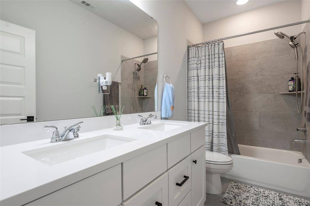 Modern bathroom featuring a double sink vanity with white cabinetry and sleek fixtures. It includes a bathtub with a gray tile surround. Well-lit and spacious, the design is both functional and stylish.