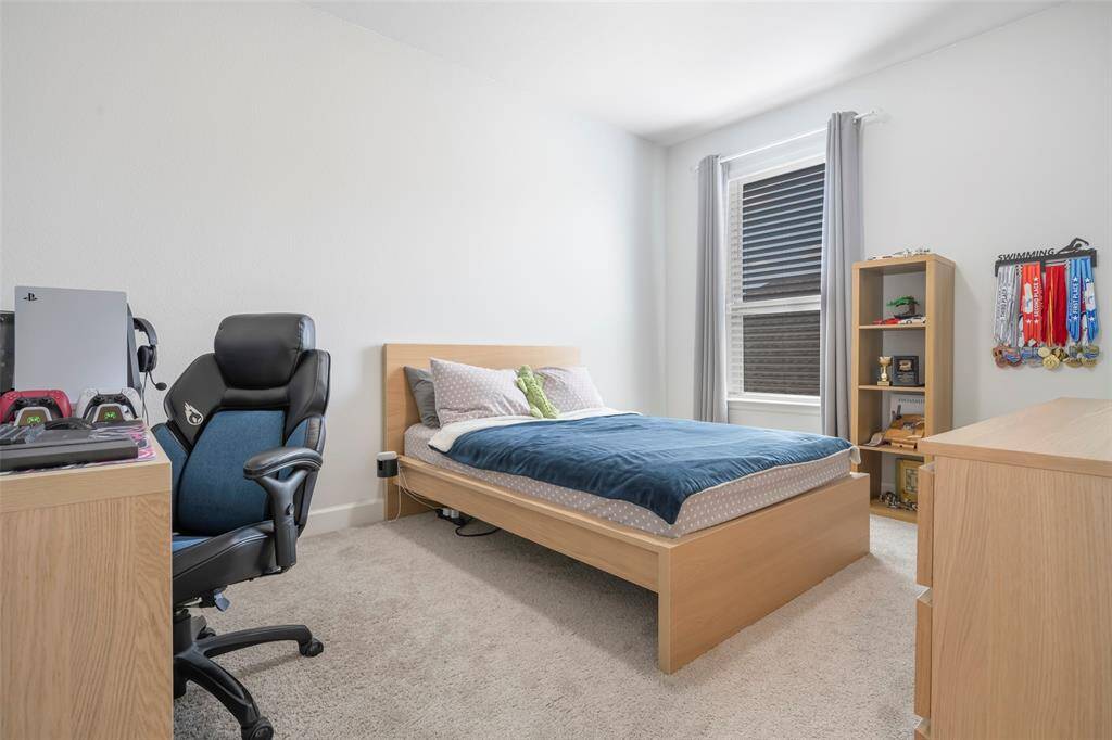 This bedroom features a cozy setup with a sleek wooden bed, a comfortable desk and gaming chair, and ample natural light from a large window.