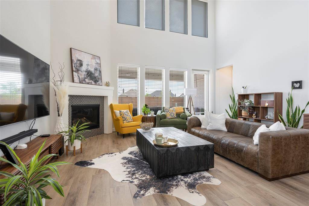 This spacious living room features high ceilings and large windows for natural light. It includes a cozy fireplace, stylish seating, and modern decor, with a wooden floor and a striking area rug. Perfect for relaxing or entertaining.