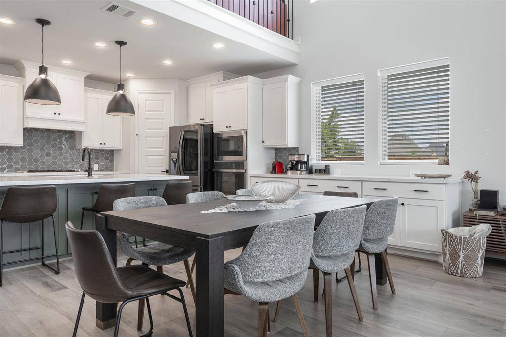 This modern kitchen and dining area features sleek cabinetry, a large island with bar seating, and contemporary pendant lighting. The neutral color palette, complemented by a hexagonal backsplash and stainless steel appliances, creates an inviting and stylish space. Large windows provide ample natural light, enhancing the open and airy feel.