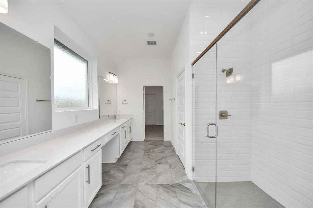 Discover a bright, modern bathroom with sleek white cabinetry, double sinks, and a spacious walk-in glass shower. Enjoy stylish tile flooring and abundant natural light from a frosted window.