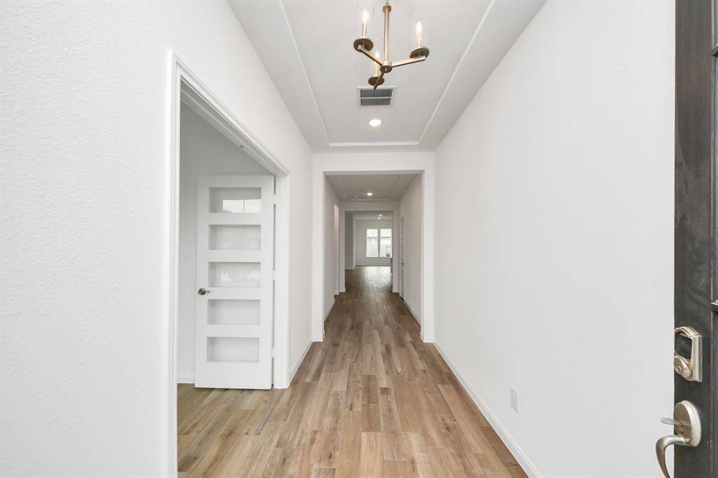 Experience a bright, modern hallway with elegant wood flooring, crisp white walls, and sophisticated recessed lighting complemented by a chic chandelier. A frosted glass door adds a touch of privacy, leading to an inviting open area.
