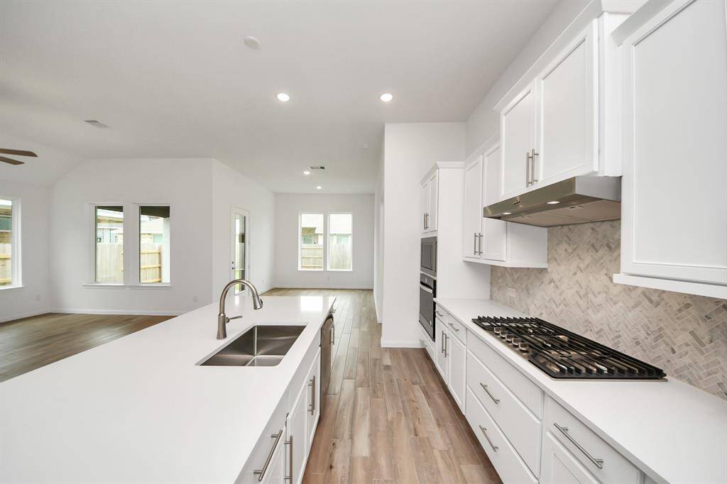 Discover an inviting, modern kitchen seamlessly blending into a bright living area. Enjoy sleek white cabinetry, a spacious island with a sink, and a gas cooktop. Large windows and wood-look flooring enhance the airy, welcoming atmosphere.