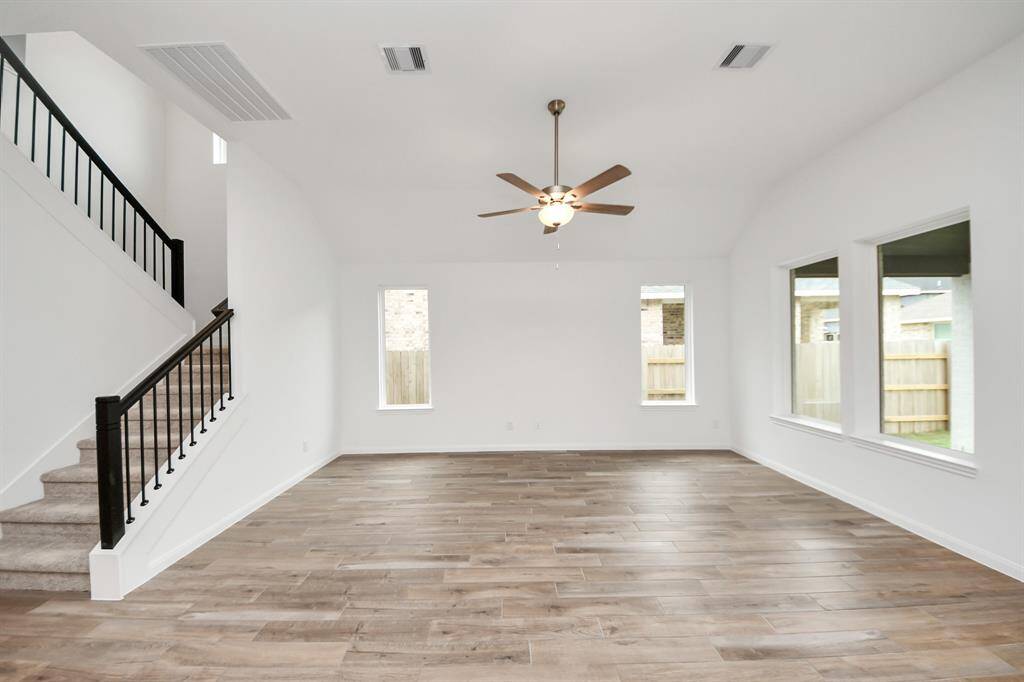 Spacious and bright living area with wood-style flooring, high ceilings, and large windows. Features a ceiling fan and a sleek staircase with dark railing, perfect for open-concept living.