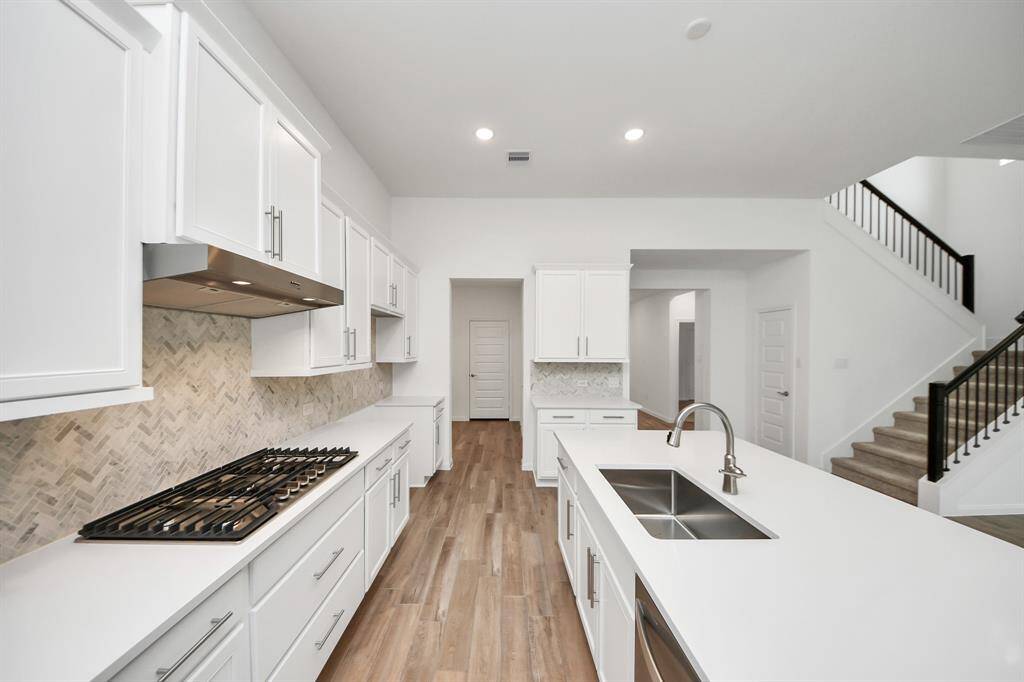 Discover a modern kitchen with sleek white cabinets, a chic herringbone backsplash, and a spacious island featuring a double sink. The open design seamlessly connects to a welcoming living area, with a staircase leading upstairs.