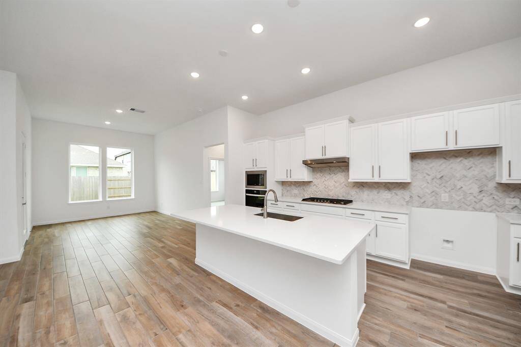 Discover this elegant kitchen featuring sleek white cabinetry, a generous island, and a chic backsplash. The open layout, enhanced by wood-style floors and abundant natural light, creates a warm and inviting atmosphere.