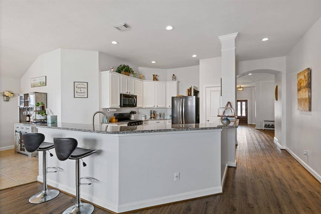 This photo showcases a modern kitchen with white cabinetry, granite countertops, and a breakfast bar with room  for seating. The open layout features hardwood floors and recessed lighting, leading to a bright hallway.