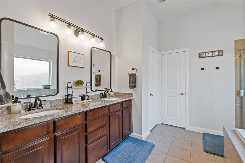 This bathroom features a double vanity with granite countertops, stylish dark wood cabinets, and modern lighting fixtures. It has a bright, clean aesthetic with light-colored walls and tiled flooring, complemented by decorative mirrors.