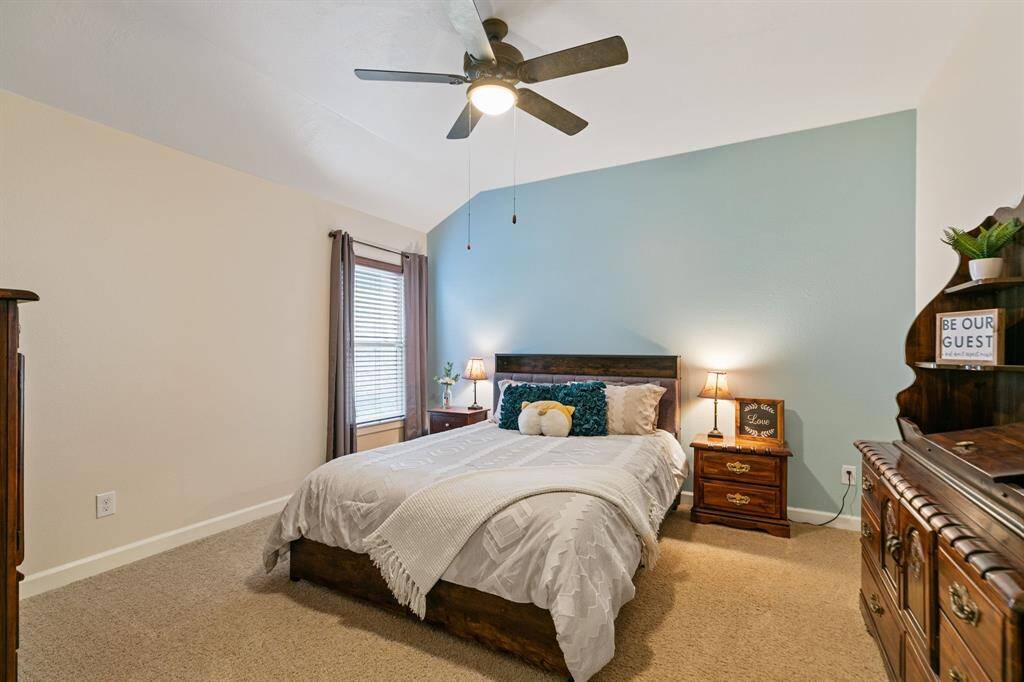 This cozy bedroom features a high ceiling with a ceiling fan, a soothing blue accent wall, and ample natural light from a large window creating a welcoming atmosphere.