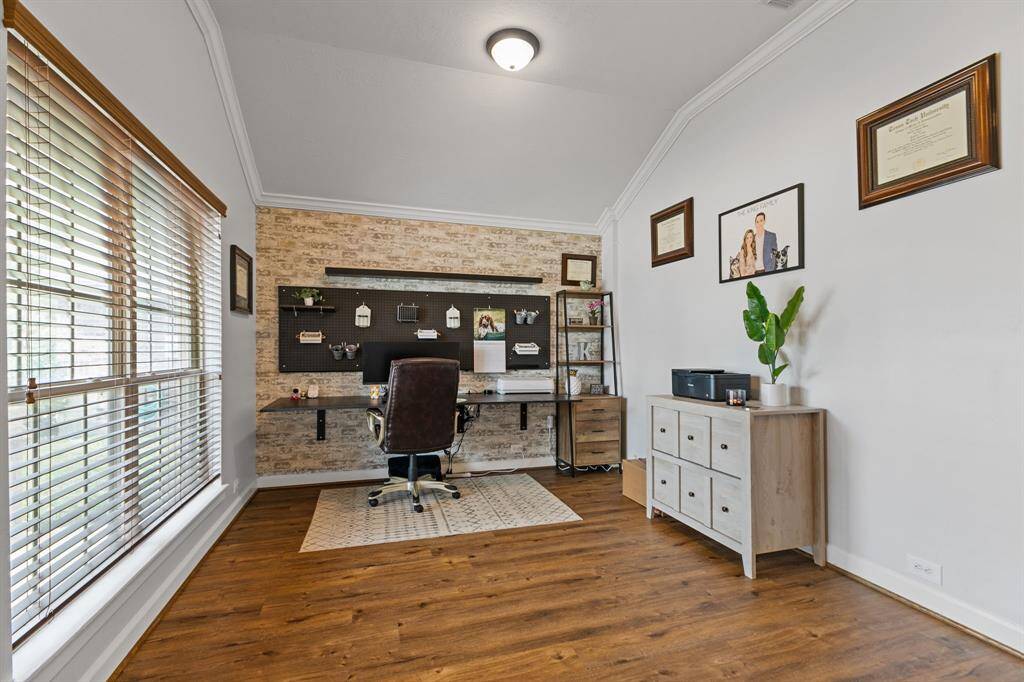 This photo showcases a bright, modern home office with a large window bringing natural light. The room features a stylish accent wall, complemented by warm laminate wood flooring and crown molding. Ideal for a functional and inviting workspace.
