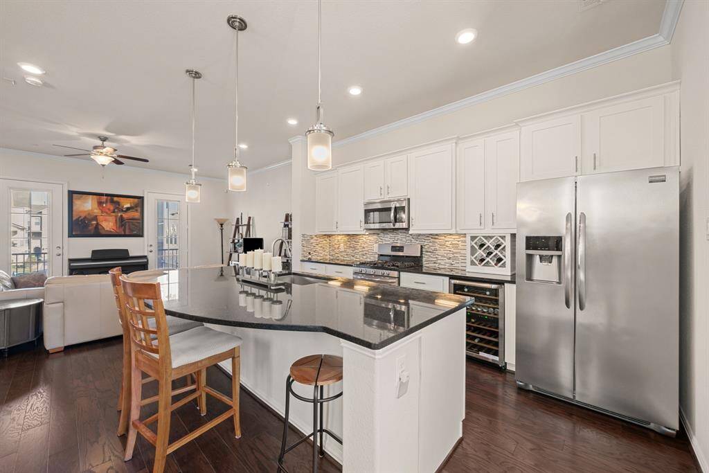 This modern kitchen features a spacious open layout with a large island, sleek granite countertops, and stainless steel appliances. It's complemented by dark hardwood floors, white cabinetry, and stylish pendant lighting, seamlessly flowing into a cozy living area with ample natural light.