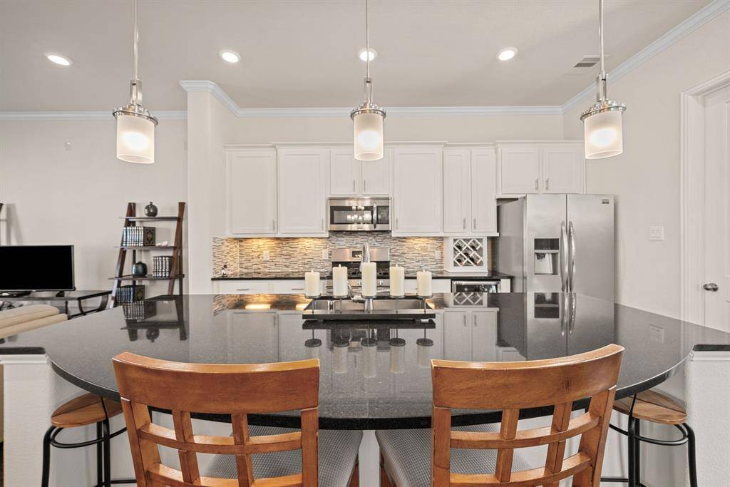 This photo showcases a spacious open-concept kitchen and dining area. The kitchen features sleek black countertops, modern stainless steel appliances, and stylish pendant lighting. The dining area includes a wooden table set under an elegant chandelier. Light wood flooring and ample natural light enhance the inviting atmosphere.