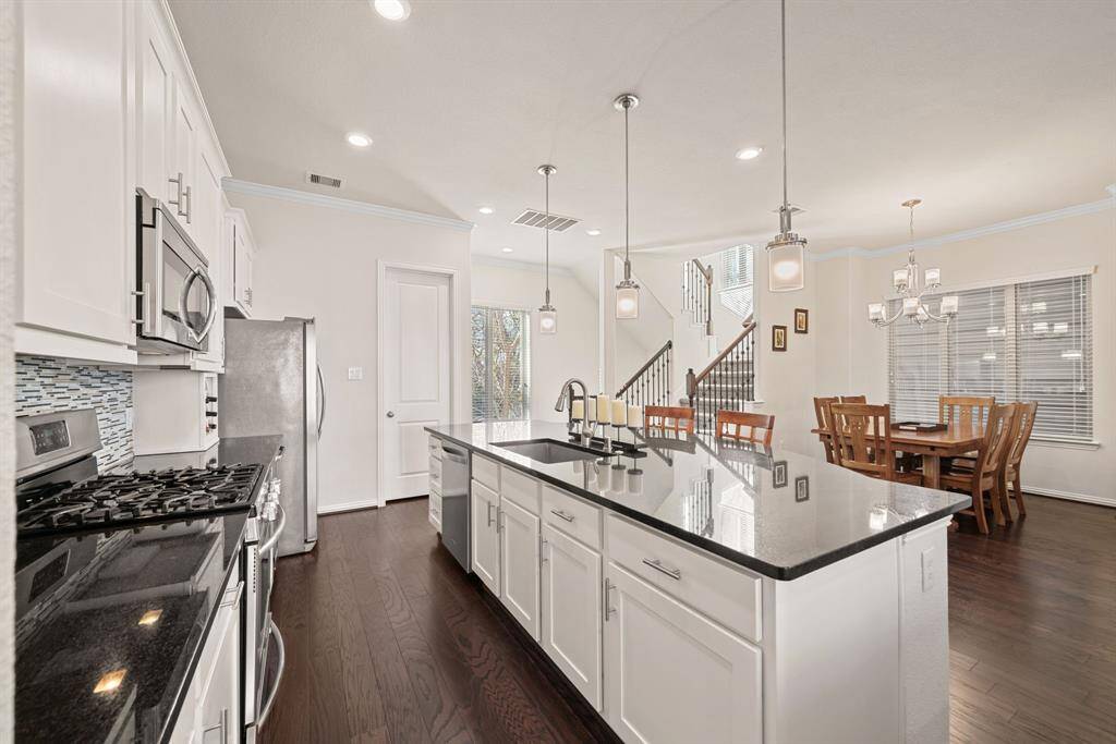 This modern kitchen features white cabinetry, a sleek black granite island with seating, stainless steel appliances, and a stylish tile backsplash. Bright and open, it also includes recessed lighting and a window for natural light.
