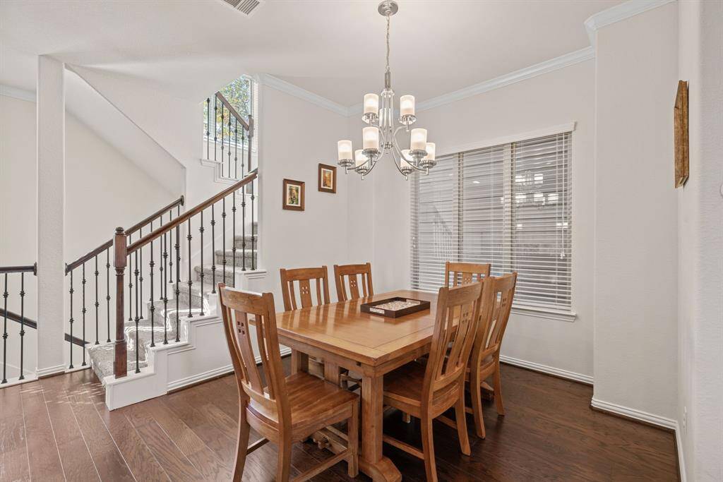 This photo showcases a bright, open-concept dining and kitchen area. The dining room features a wooden table with matching chairs under a stylish chandelier. The kitchen includes modern stainless steel appliances, white cabinetry, and a breakfast bar with pendant lighting. A staircase with decorative railing adds elegance to the space.