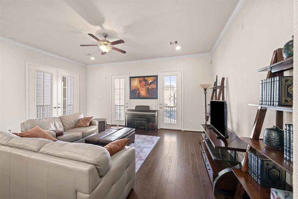 This spacious living room features elegant hardwood floors, a ceiling fan, and ample natural light from multiple glass doors. It is tastefully furnished with white sofas, a TV, and modern shelving, creating a comfortable and inviting atmosphere.