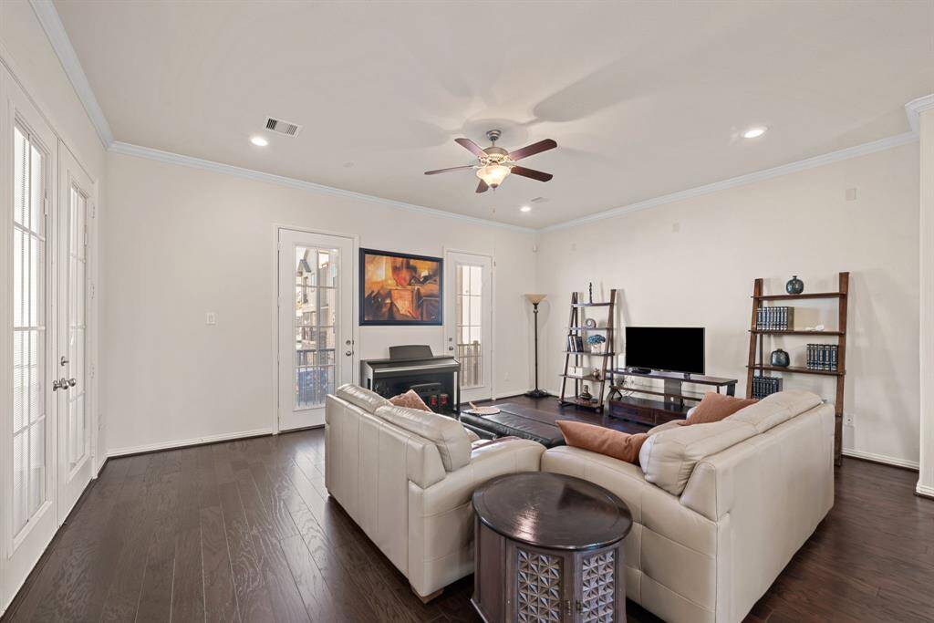 This open-concept living space features hardwood floors, a cozy sectional sofa, and a modern kitchen with white cabinetry and stainless steel appliances. The room is well-lit with recessed lighting and a ceiling fan, and offers a comfortable, inviting atmosphere with large windows providing natural light.