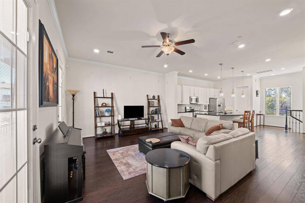 This open-concept living area features a cozy seating arrangement with a sectional sofa, a central ottoman, and a piano. The space is well-lit with recessed lighting, a ceiling fan, and large windows. The modern kitchen includes pendant lights, an island, and adjacent dining area. Dark wood floors add warmth throughout.
