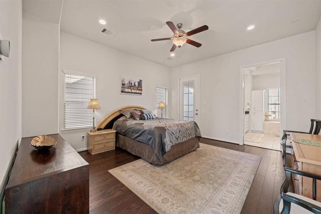 This spacious bedroom features hardwood floors, a ceiling fan, and neutral walls. It includes a cozy area with a dresser and mirror, plus two chairs. An open doorway leads to a bathroom, enhancing convenience and accessibility.
