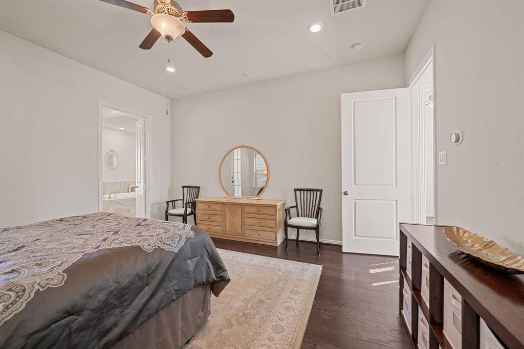 This spacious bedroom features elegant hardwood flooring, a ceiling fan, and ample natural light. It includes a stylish dresser with a round mirror, accent chairs, and a cozy bed with patterned bedding. The room connects to another area through a double door, adding to its appeal.