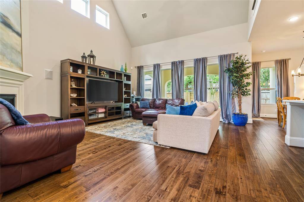 Spacious living room with high ceilings, large windows, and abundant natural light. Features wood flooring, a cozy seating area, and an entertainment center. Adjacent to an open dining area and kitchen, perfect for family gatherings.